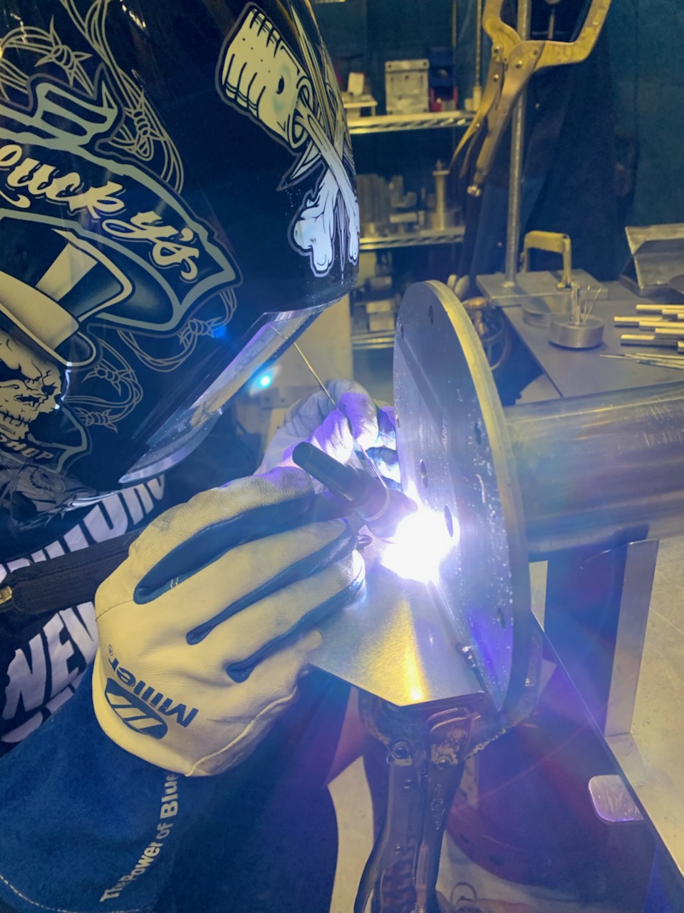 Welder TIG welding a circular steel flange on a workshop bench, the arc lighting the work